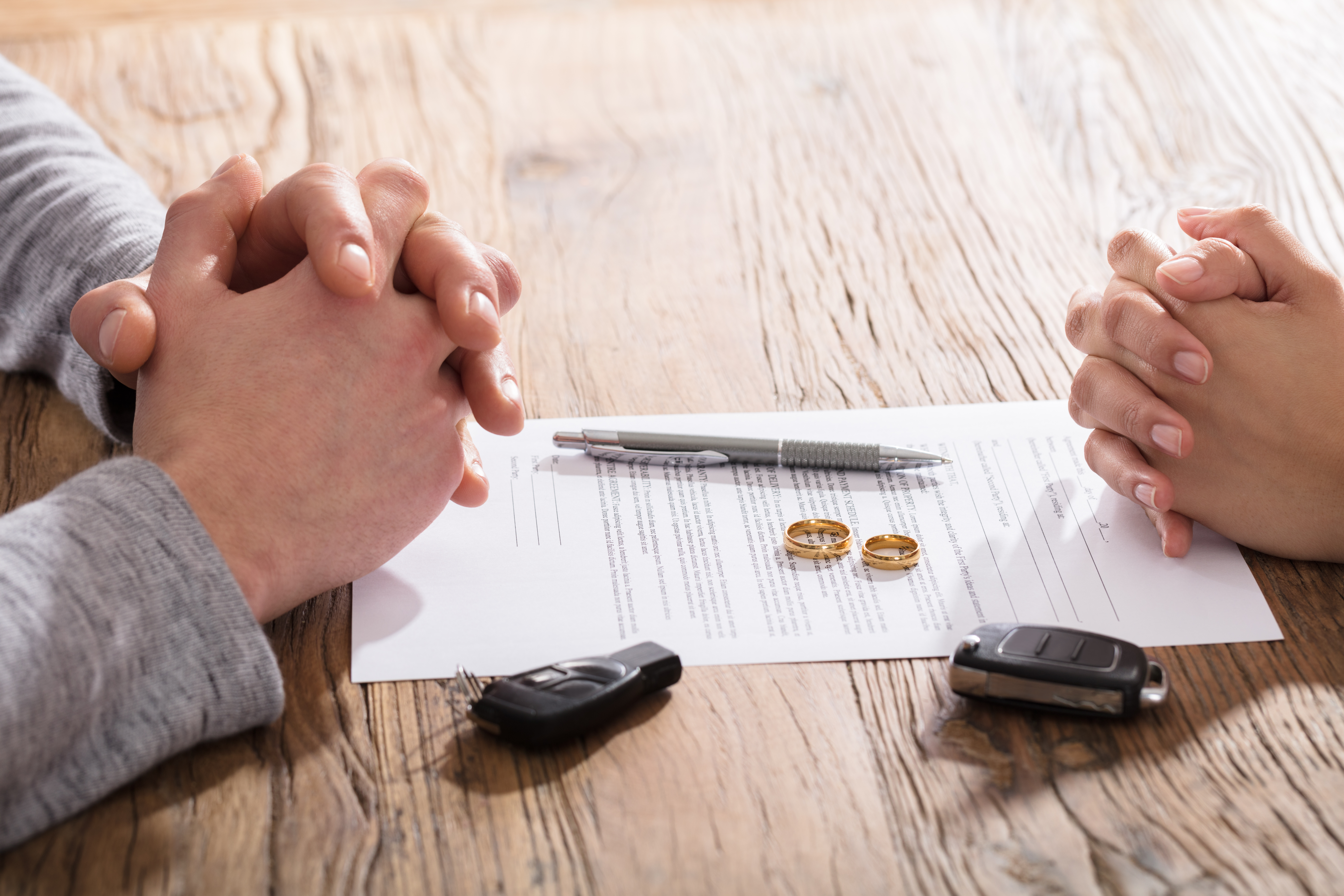 Two pairs of clasped hands are seen across a wooden table with a legal document in between. Two gold rings, a car key, and a pen rest on the paper. The scene suggests a negotiation or agreement process.