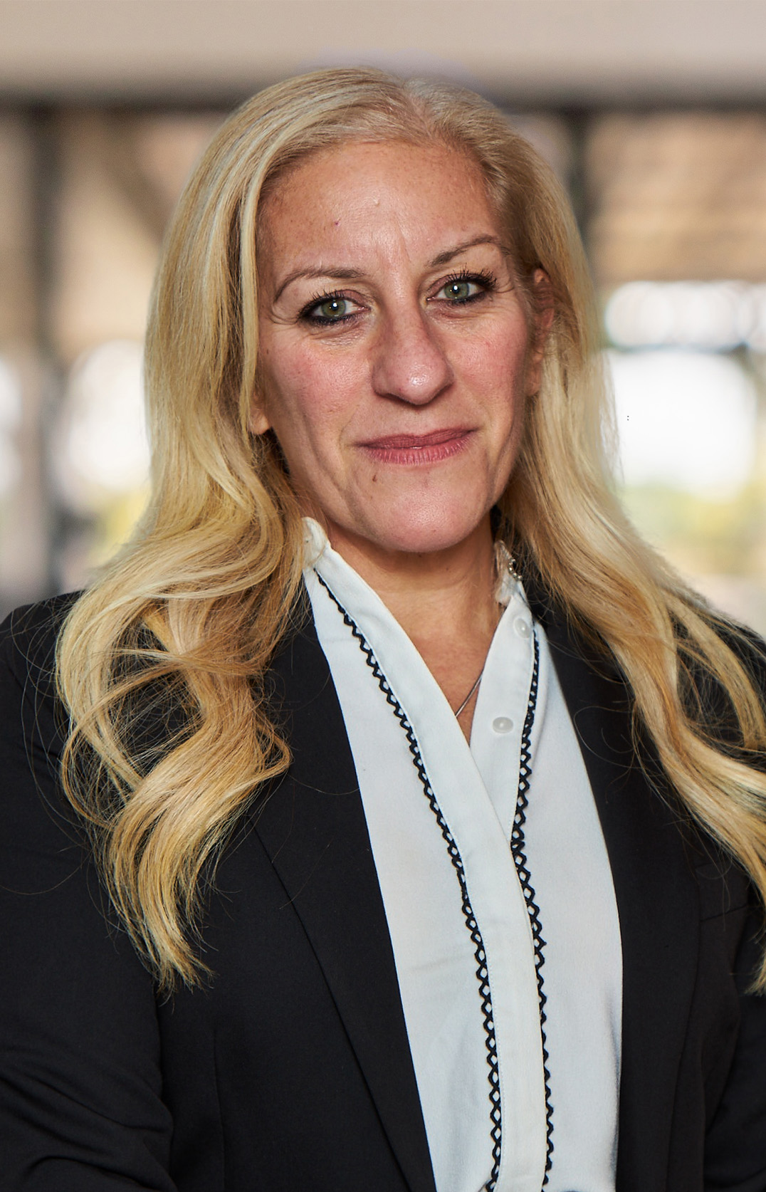 Anna Maria Petracca, a woman with long blonde hair, wears a black blazer over a white blouse. She poses with a neutral expression in an indoor setting, surrounded by blurred elements in the background.