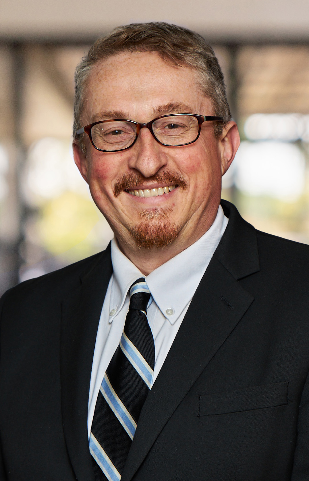 Eric A. Parisi, a man with short hair, glasses, and a mustache, is smiling warmly. He is dressed in a black suit, white shirt, and striped tie. The softly blurred background hints at an indoor setting.