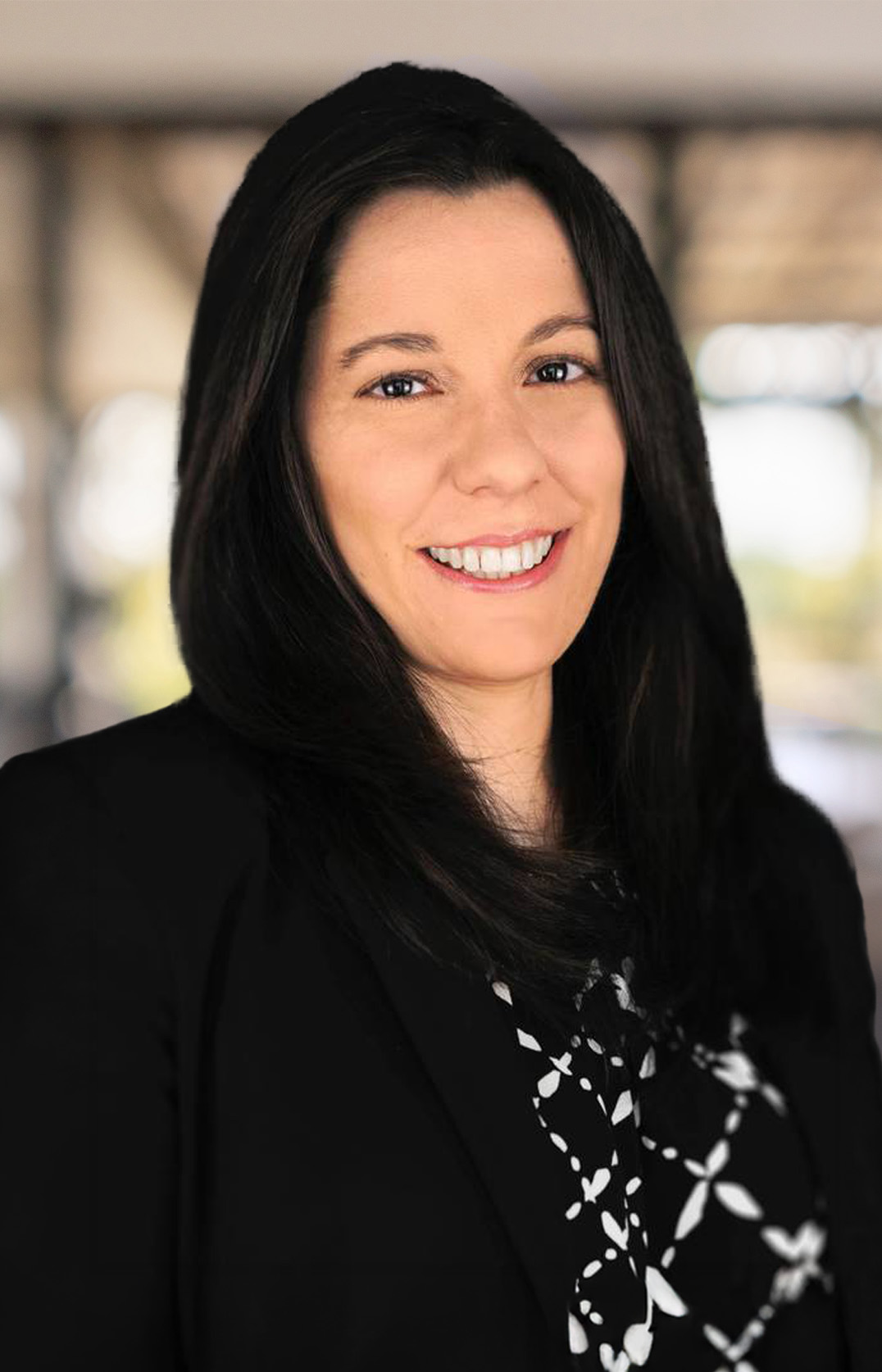 Cynthia J. Lambo, a woman with long dark hair, smiles at the camera in a black blazer over a patterned blouse. The softly blurred background hints at an indoor setting, adding to her poised and professional demeanor.